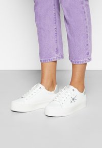Baskets blanches en matériau lisse, à bout rond et avec des accents argentés au niveau du talon. Elles présentent des lacets et un logo sur le côté. Portées avec un jean cropped lavande.
