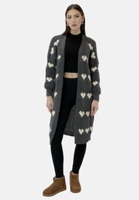 Cardigan grigio lavorato a maglia con motivi a cuore bianchi, apertura frontale e polsini a coste. Abbinato a top nero, leggings e stivali marroni alla caviglia.