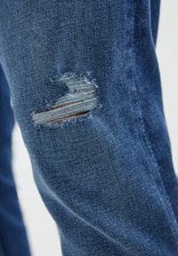 Vaqueros de mezclilla azul con un parche desgastado que presenta bordes deshilachados, exponiendo hilos horizontales y verticales en un tono azul más claro.