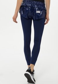 Person klädd i marinblå leggings, blanka marinblå shorts med Adidas-logotyp och svarta och grå löparskor som står mot en vit bakgrund.