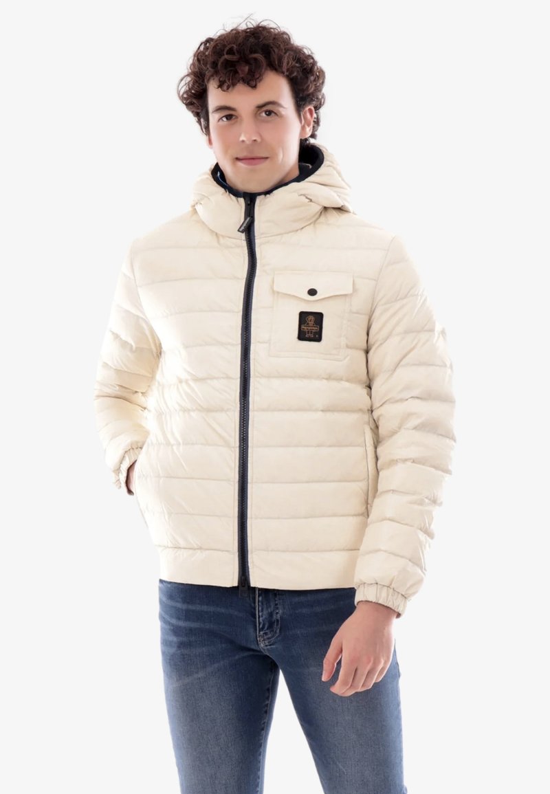 Giacca imbottita di colore crema con chiusura a zip, cappuccio e polsini a costine. Presenta una tasca sul petto con un'etichetta nera e un design strutturato.