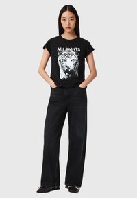 Tricou grafic negru cu imprimeu de tigru, purtat cu blugi negri cu talie înaltă și crăpătură largă. Modelul stă cu mâinile în buzunare.