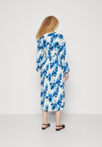 Langermet blå blomstermønstrete kjole med innsnevring i midjen og A-linje silhuett, laget av lett stoff. Svarte sandaler fullfører antrekket.