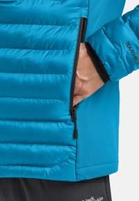Giacca isolante blu con texture trapuntata e zip nera. Presenta una tasca con zip e design dei polsini per calore e comfort.