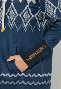Navyblauwe hoodie met witte geometrische patronen, trekkoord met oranje uiteinde, en een contrasterende zwarte manchette met oranje tekst.