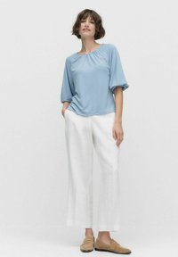 Lys blå bluse med rynket halsudskæring, løs pasform og korte pufærmer, stylet med hvide linnedbukser og brune loafers.