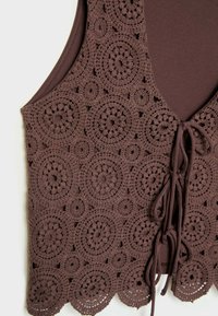 Brązowy haftowany kamizelka z kwiatowymi wzorami, w oversize'owym fasonie z wiązaniem z przodu i falbaniastym dołem. Gładka wewnętrzna tkanina kontrastuje z teksturowanym zewnętrznym wzorem szydełkowym.