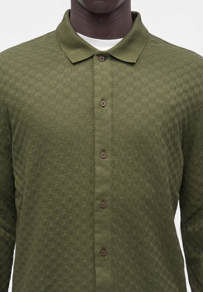 Homme portant une chemise texturée vert olive à boutons avec col par-dessus un sous-vêtement blanc uni, visible du cou à la poitrine.