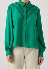 Blusa verde con bottoni, decorata con pizzo sulle spalle, maniche lunghe con polsini arricciati e vestibilità rilassata. Tessuto morbido e leggero.
