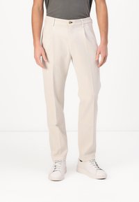Lichtbeige pantalon met een rechte pijpdesign en lopende plooien aan de voorkant. Voorzien van een knoopsluiting en riemlussen, gecombineerd met witte sneakers.