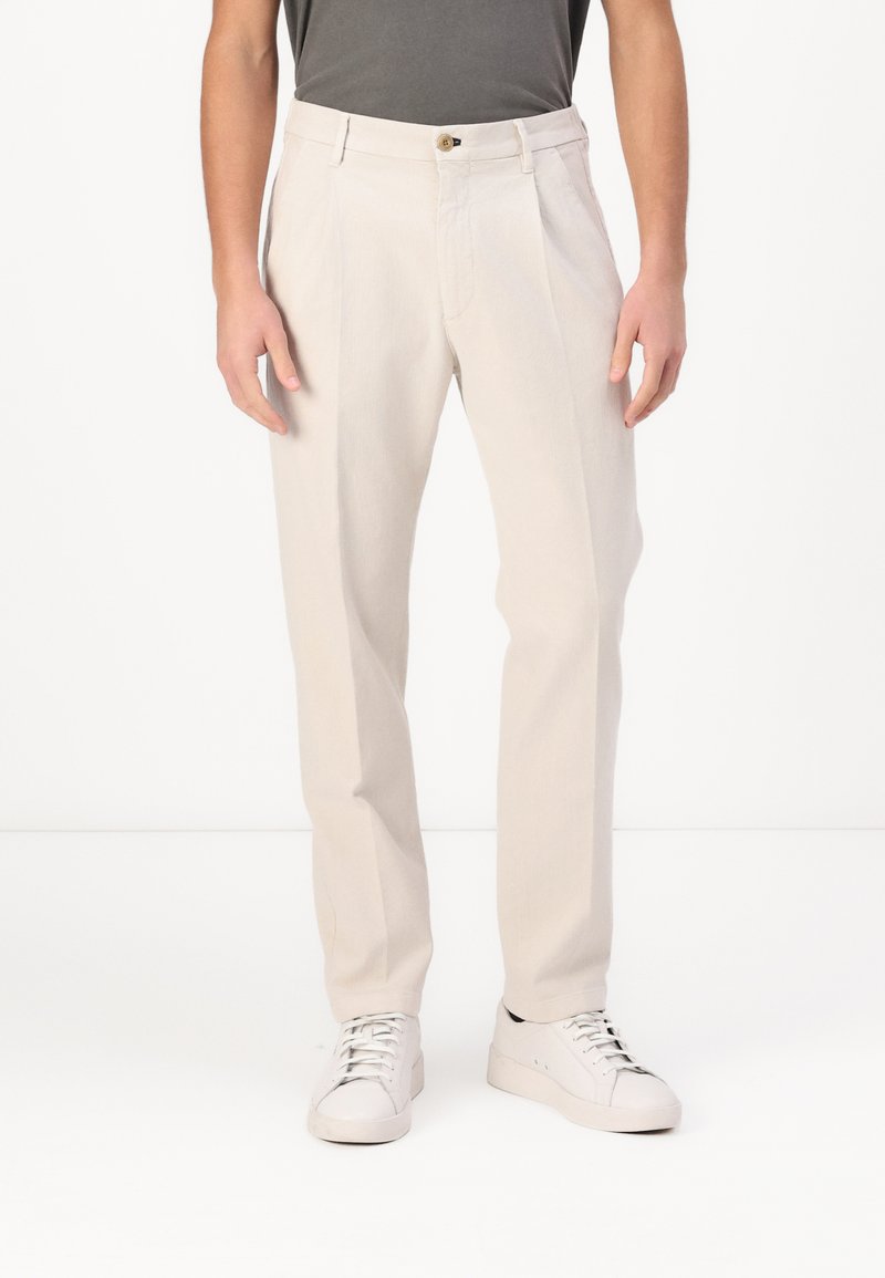 Lichtbeige pantalon met een rechte pijpdesign en lopende plooien aan de voorkant. Voorzien van een knoopsluiting en riemlussen, gecombineerd met witte sneakers.
