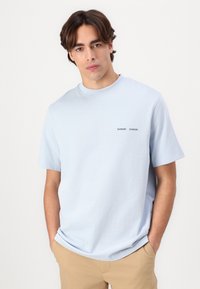 Tricou scurt cu mânecă, de culoare bleu deschis, realizat din bumbac moale. Prezintă un logo mic cu textul „SAMSOE” în albastru închis pe piept. Design cu o croială lejeră.