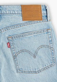 Light blue denim jeans feature a tan leather patch with embossed logo, orange stitching, and a signature pocket design with a red tab.