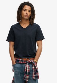 Donkerblauwe V-hals t-shirt van zacht materiaal, gecombineerd met blauwe jeans en een rood geruit shirt om de taille geknoopt. Een bruine riem en horloge accentueren de look.