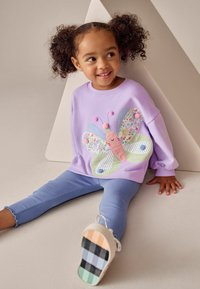 Bluză lavandă cu un design cu fluture care include modele florale și dungi, asortată cu colanți albastru deschis cu rețea și pantofi multicolori.