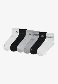 Six pairs of socks in white, black, and gray, featuring a green Lacoste logo. Each pair has a ribbed cuff and smooth texture.