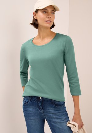 Top verde menta de manga larga con escote redondo y mangas de 3/4, combinado con jeans de mezclilla azul. Textura suave, diseño informal.