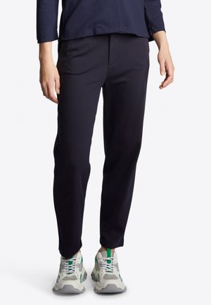 Navy trousers made of a smooth fabric, featuring a tapered fit, two front pockets, and paired with gray and green athletic sneakers.