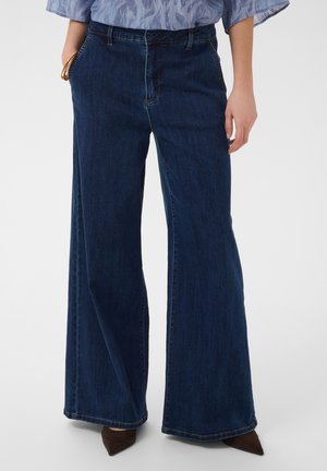 Frau trägt dunkelblaue Jeans mit weitem Bein, eine hellblaue gemusterte Bluse, die in die Hose gesteckt ist, und braune spitze Schuhe.