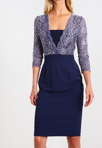 Femme portant une robe bleue marine longueur genou avec un haut à motif en dentelle, manches trois-quarts et un décolleté plongeant en V sur un sous-vêtement uni.