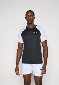 T-shirt de sport noir et blanc à manches courtes, avec une texture lisse et un logo sur la poitrine. Porté avec un short blanc.