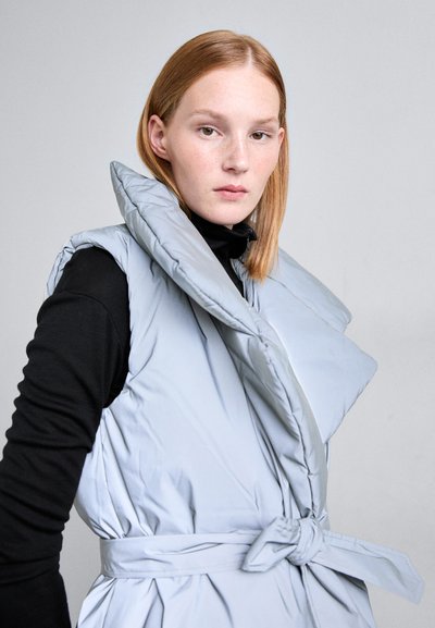 Ljusblå, ärmlös pufferväst med en stor krage, midje-knut och slät, vadderad textur. Stylad över en svart topp.