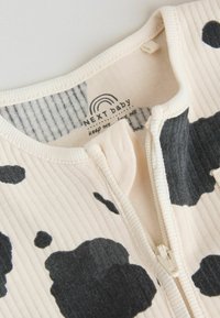 Elfenbeinfarbener Babystrampler mit Rippenstruktur, schwarzem Kuhmuster, Frontreißverschluss und gestreiftem Innenfutter. Weicher Baumwollstoff für Komfort.