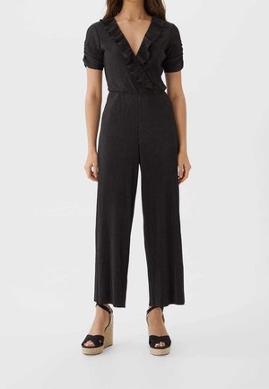 Vrouw draagt een zwarte geribbelde jumpsuit met korte mouwen, ruches aan de V-hals, broek met wijde pijpen en zwarte sandalen met sleehak.