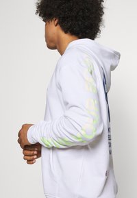 Vit hoodie med avslappnad passform, med stora gröna och blå bokstäver på framsidan och ner längs ena ärmen. Mjukt tyg, dragsko i huvan.