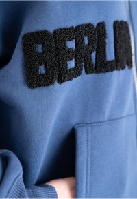 Hoodie blu con lettering "BERLIN" testurizzato in nero, tasca a marsupio, polsini a costine e un tessuto in cotone morbido.