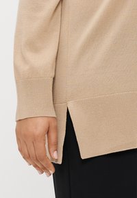 Μπεζ πλεκτό πουλόβερ με ριμπ μανσέτες και στρίφωμα, με πλαϊνές σχισμές. Μαλακή υφή με άνετη γραμμή, παρουσιάζεται με ένα χέρι πάνω στο ύφασμα.