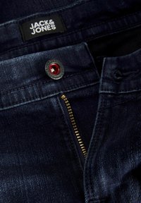 Tumed sinise denimi püksid, millel on hõbedane tõmblukk, punane nupp ja must silt "JACK & JONES" vöökoha sees.