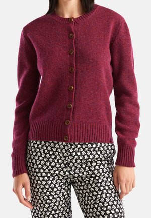 Kobieta ubrana w zapinany na guziki bordowy cardigan z wełny i czarno-białe spodnie w kwiatowy wzór, stojąca z rękami swobodnie opuszczonymi wzdłuż ciała.