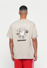 Beige bomulds t-shirt med et cartoon-illustration af en iskold latte på ryggen, med teksten "ADIDAS" og "ORIGINALS" i kontrastfarver.