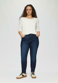 Biała koszula z długim rękawem, ciemnoniebieskie dżinsy i beżowe trampki z czarnymi akcentami. Model stoi z rękami w kieszeniach na jasnym tle.