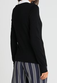 Maglione nero a maniche lunghe con un design attillato e polsini a coste, abbinato a pantaloni a righe blu e bianche, che mostrano un look informale a strati.
