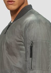 Graue Bomberjacke aus leichtem Material, mit geripptem Kragen, einem Reißverschlussfach am Arm und matter Oberfläche.