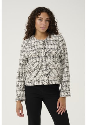 Korte jas in een zwart-wit geruit patroon met textuurdetails, ronde halslijn en voorzakken. Gestyled met zwarte broek.