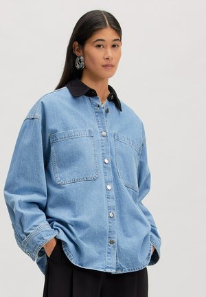 Femme portant une veste en jean bleu clair oversize avec col noir et grands anneaux en argent, mains dans les poches, fond neutre.