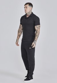 Camisa polo negra de manga corta, fabricada en tejido texturizado, combinada con pantalones jogger negros que presentan un pequeño logotipo. Zapatillas deportivas negras.