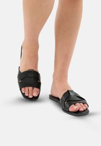 Sorte lædersandaler med flad sål, udstyret med krydsrem-design, åbne tæer og subtile hardware-detaljer.