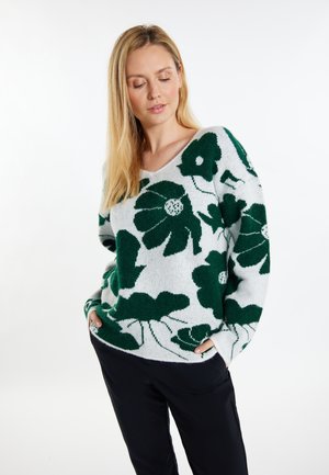 Sweter dzianinowy o luźnym kroju, z białym tłem i zielonym wzorem kwiatowym. Wykończenie w serek oraz ściągacze. Materiał wydaje się miękki i teksturowany.