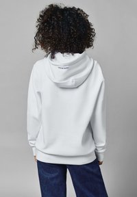 Sudadera blanca con un bolsillo tipo canguro, puños y dobladillo acanalados. Presenta un diseño simple con una marca visible en el forro interior de la capucha.