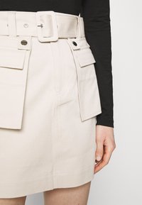 Detailní záběr na béžovou mini sukni s ladícím páskem a kapsami na klopě, která je kombinována s černým topem s dlouhým rukávem, na fotografii je vidět uvolněná pravá ruka.