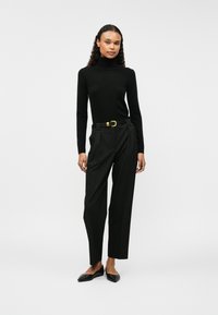 Μαύρο πουλόβερ με ζιβάγκο συνδυασμένο με μαύρους ψηλόμεσους πλισέ παν trousers, που ολοκληρώνεται με μια ζώνη με χρυσό πόρπη και μαύρες μυτερές μπαλαρίνες.