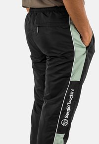 Zwarte en mintgroene trainingsbroek met witte piping en het "Sergio Tacchini"-logo aan de zijkant, gedragen door een persoon met handen in de zakken.