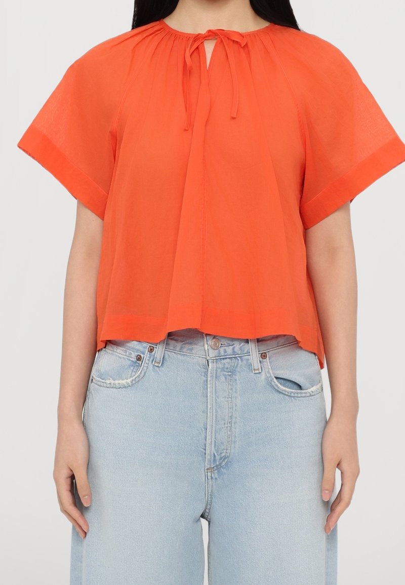 Femme portant un chemisier orange ample à manches courtes avec un noeud au col et un jean taille haute bleu clair.