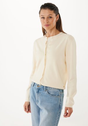 Žena s světle hnědými vlasy, oblékající krémový cardigan na knoflíky a světle modré džíny, stojící na bílém pozadí.