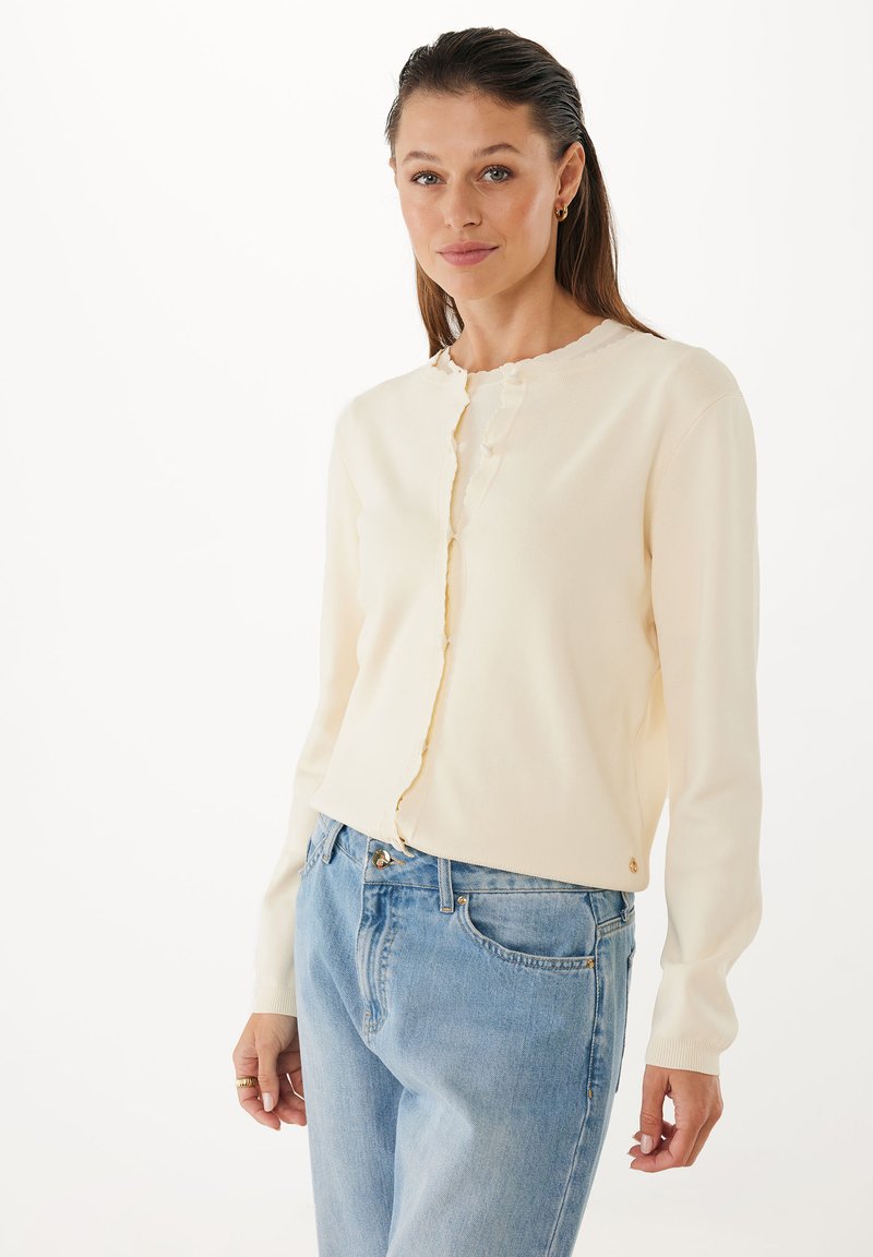 Kvinna med ljusbrunt hår iförd en krämfärgad knäppt cardigan och ljusblå jeans, stående mot en vit bakgrund.