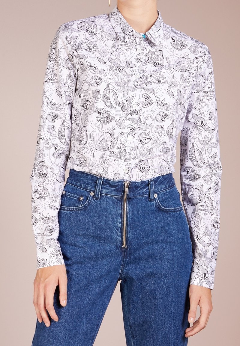 Camicia bianca a maniche lunghe con intricate fantasie floreali e animali nere, abbinata a jeans blu a vita alta con zip frontale.